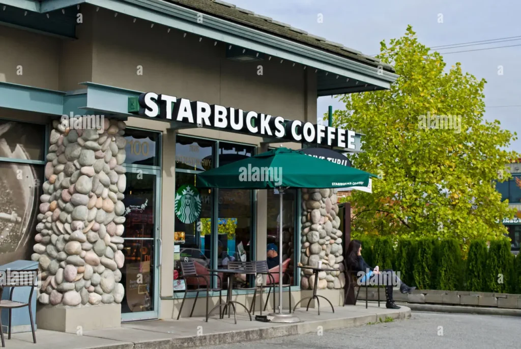 Exterior view of a Starbucks coffee shop showcasing the storefront sign, stone pillars, and outdoor seating area with a branded umbrella