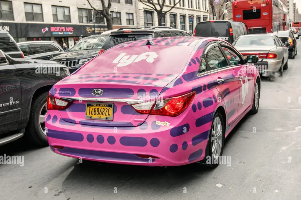 A Lyft-branded pink and purple Hyundai sedan driving in New York City displaying the distinctive rideshare design